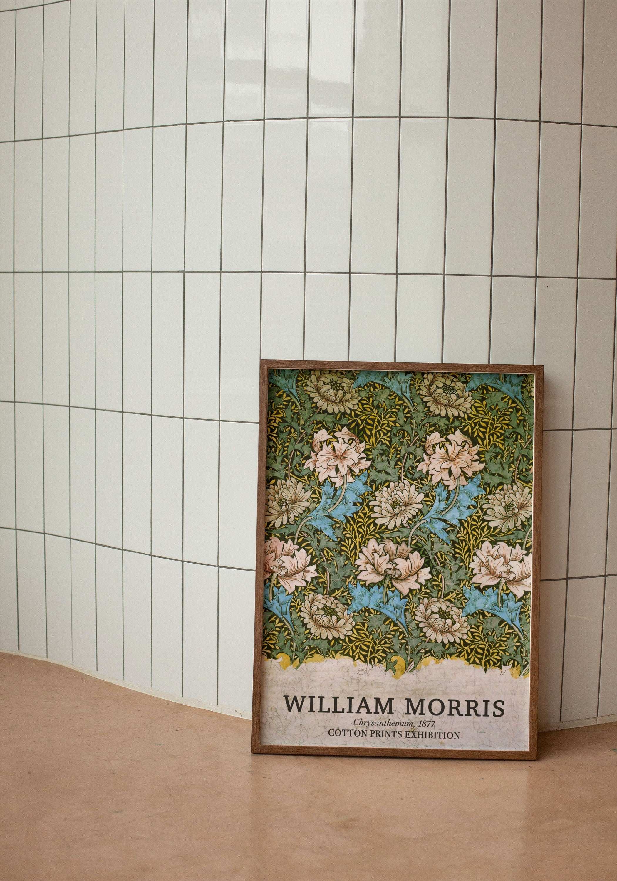 Modern beyaz dikey fayanslı bir duvarın önünde, yerde duran doğal ince ahşap çerçeveli William Morris Chrysanthemum 1877 çiçek desenli sergi posteri