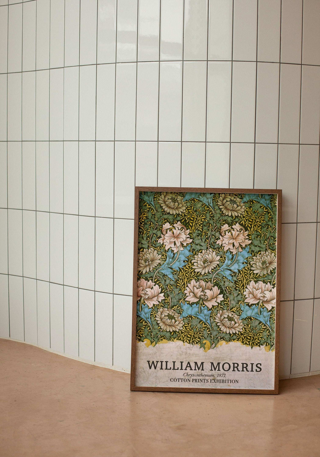 Modern beyaz dikey fayanslı bir duvarın önünde, yerde duran doğal ince ahşap çerçeveli William Morris Chrysanthemum 1877 çiçek desenli sergi posteri