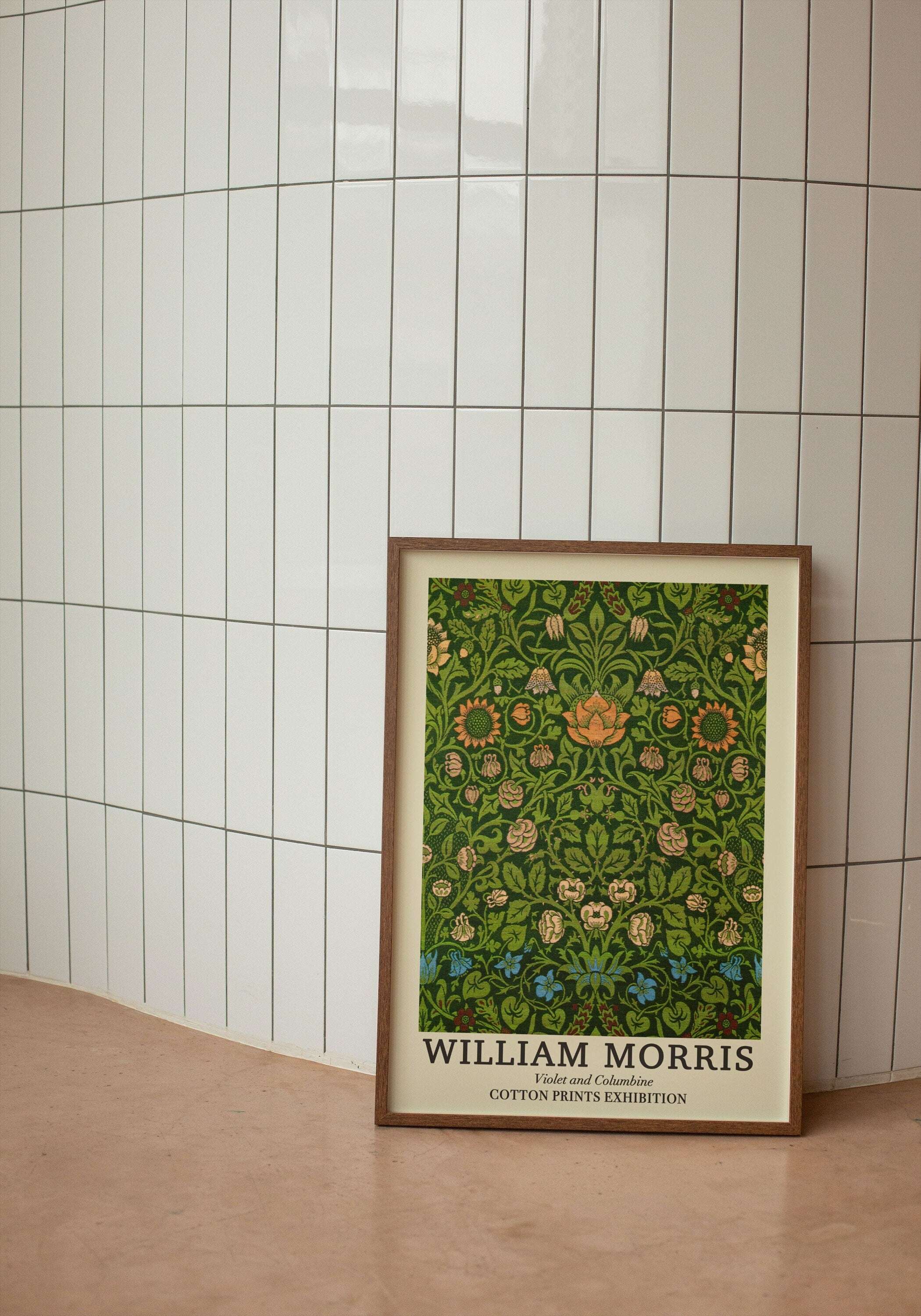 Beyaz dikey seramik duvar önünde yerde duran, ahşap çerçeveli William Morris Larkspur 1875 botanik desenli sergi posteri