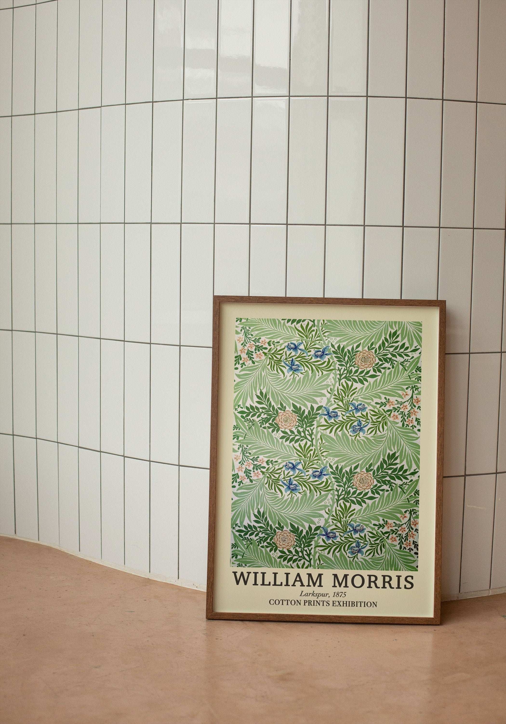 William Morris Larkspur 1875 tasarımı yeşil botanik desenli sanat posteri, ahşap çerçeve içerisinde beyaz dikey fayanslı duvarın önünde yerde duruyor.